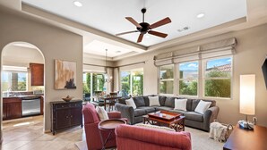 A spacious living area with a gray sofa, red armchairs, and a dining table. A ceiling fan and large windows provide natural light. - La Quinta Resort-Style Retreat (La Quinta)