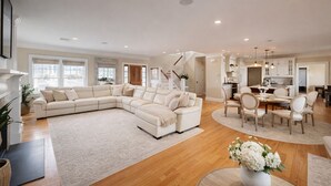 A spacious living room with a large L-shaped sectional sofa, a dining area with a round table, and a kitchen in the background.