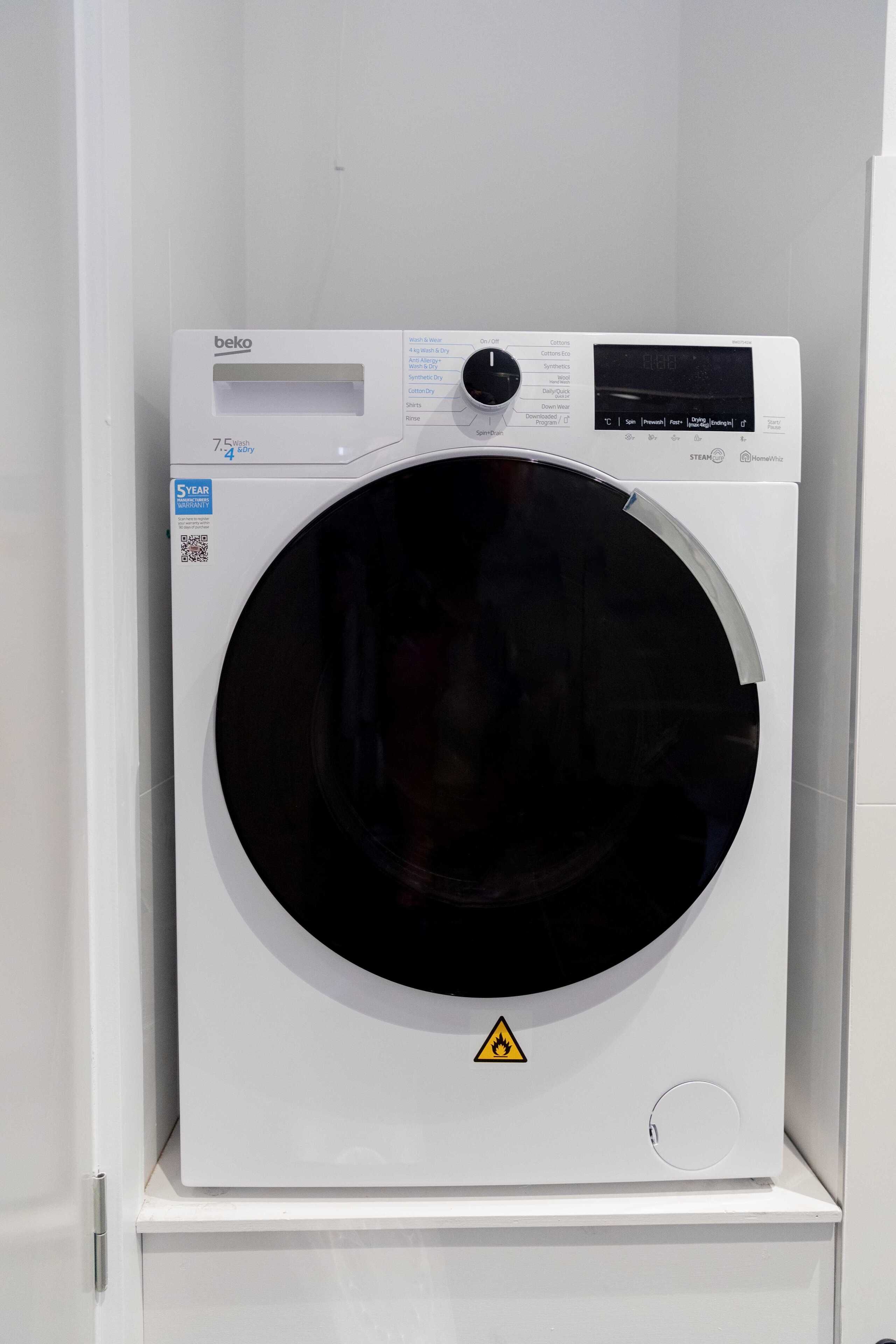 A white wall-mounted washing machine with a digital display, a control panel, and a safety warning symbol.