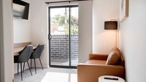 A modern room with a sliding glass door leading to a balcony, a sofa, a desk with two chairs, and a remote control on a side table. - Amica Serviced Apartments (Orange)