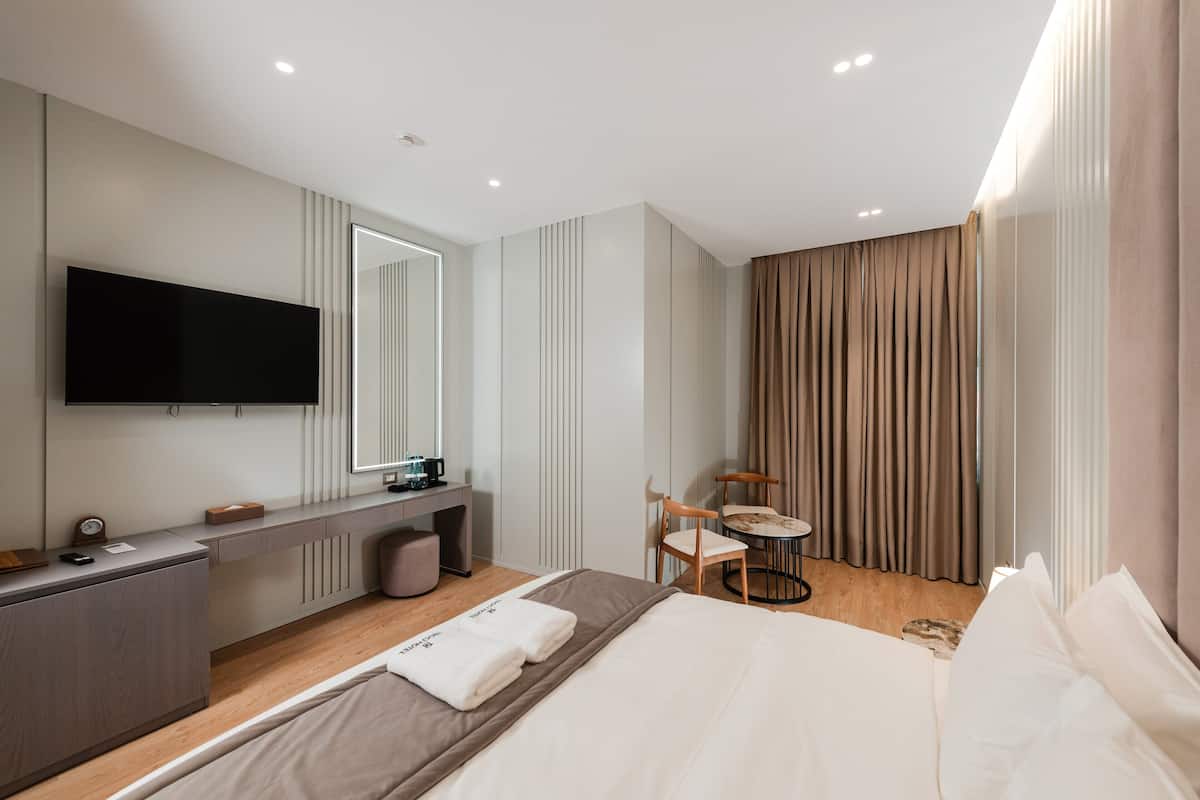 A modern hotel room with a large bed, a desk with a flat-screen TV, a small round table, and a chair.