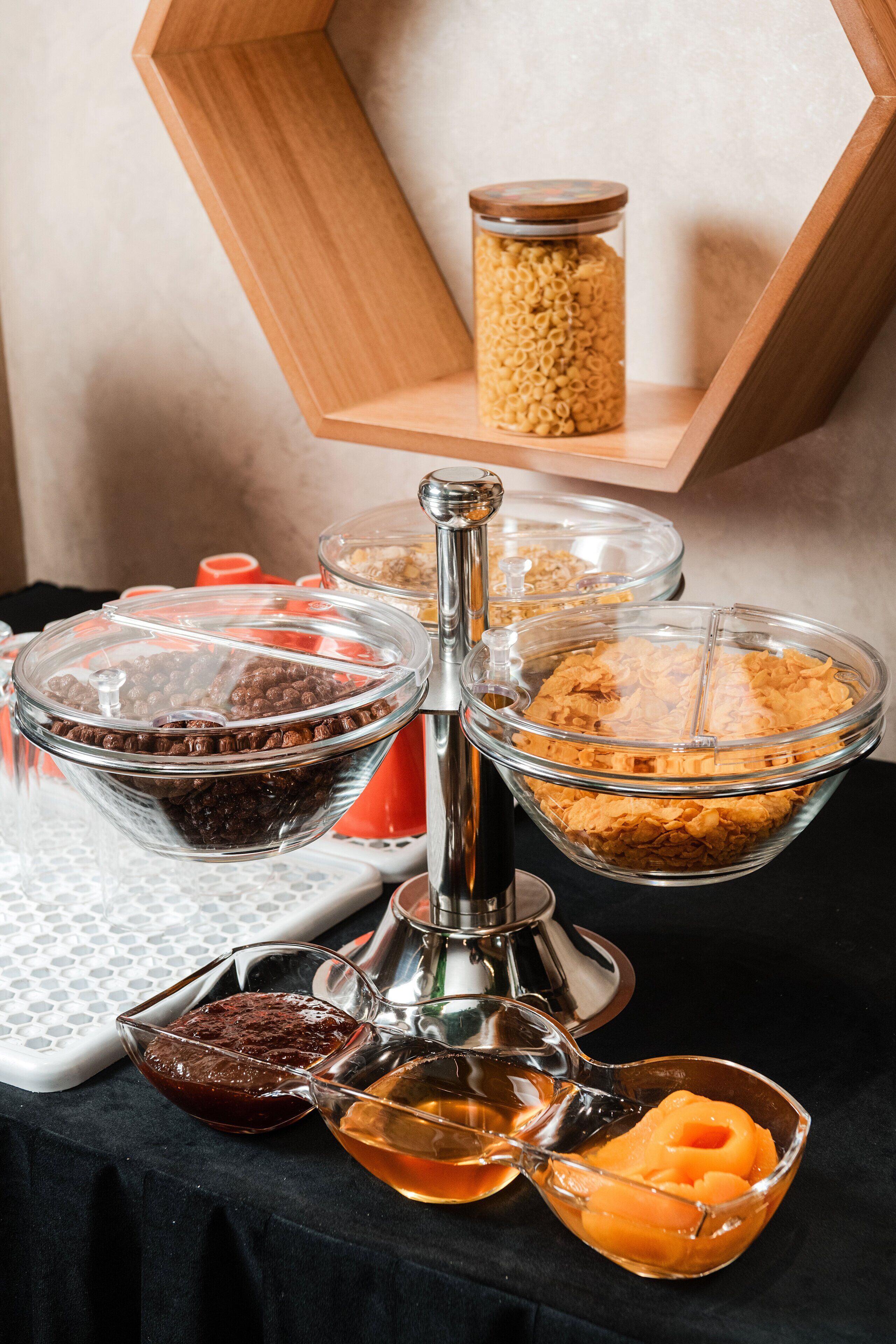 Een buffetopstelling met diverse soorten eten in glazen schalen en bakken, een houten plank met een pot, en een metalen doseerder.