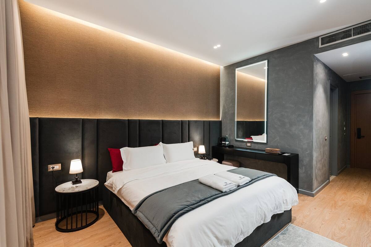 A modern hotel room with a large bed, bedside tables, a mirror, and a dark headboard.
