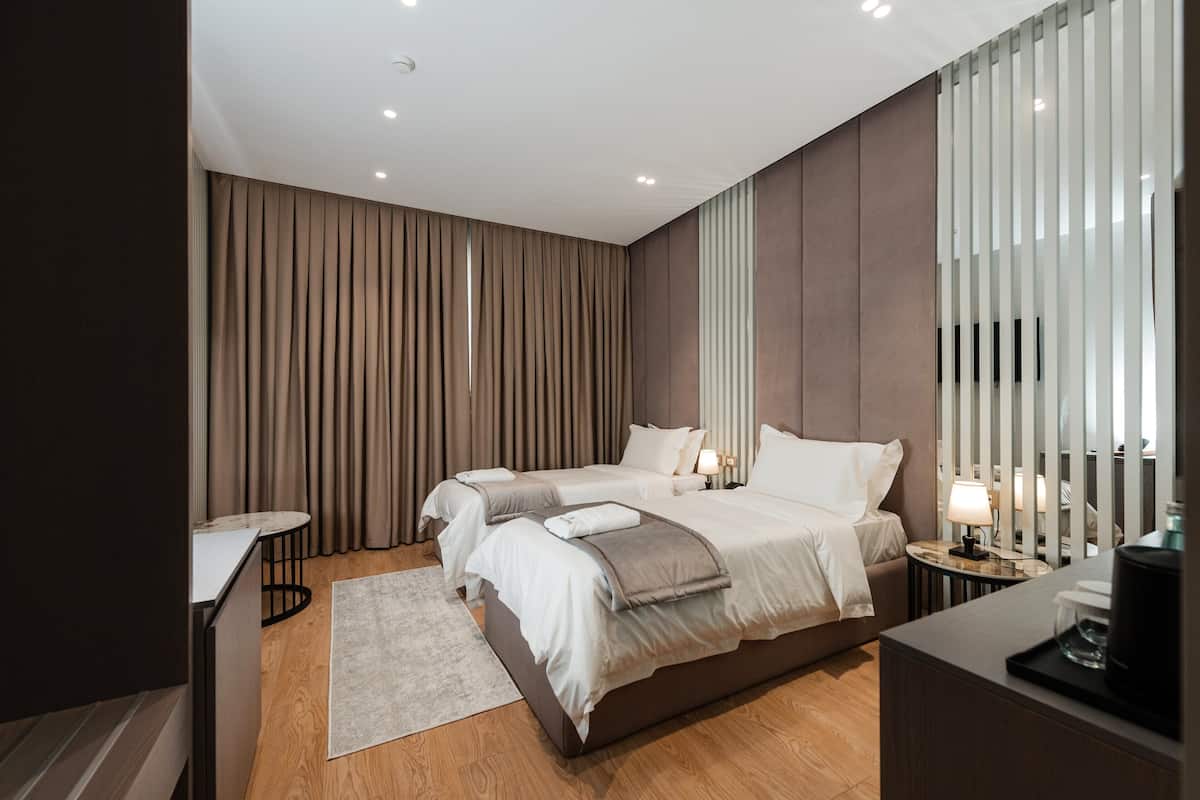 A modern hotel room with two beds, a small table, a bedside table with a lamp, and a large window with blinds.