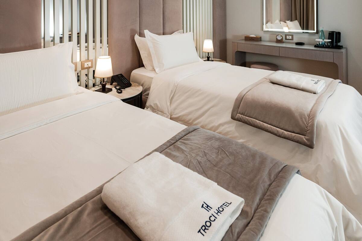 A hotel room with two beds, a bedside table with a lamp, a desk with a mirror, and a wall-mounted light fixture.