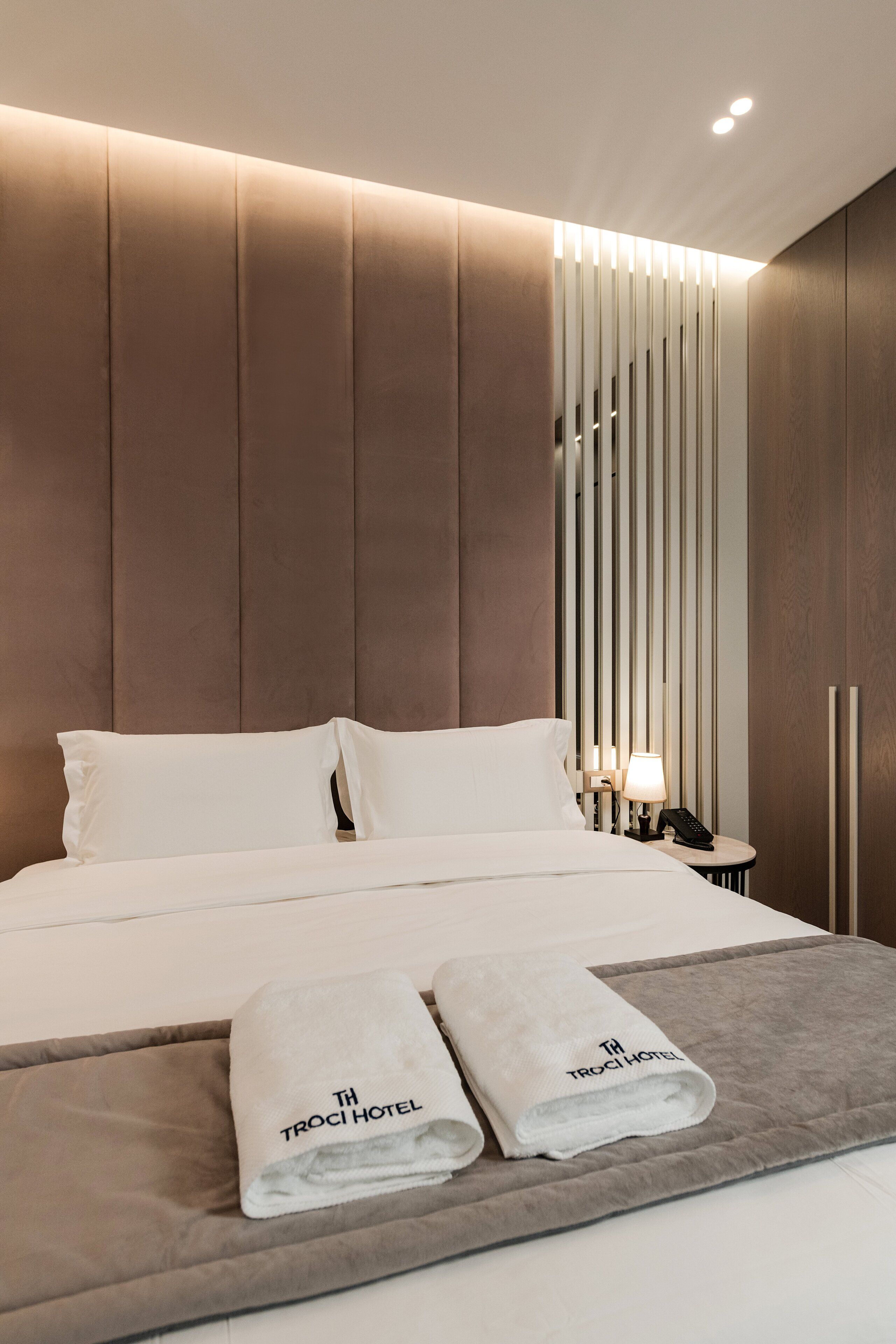 A modern hotel room with a large bed, two pillows, a bedside table with a lamp, and two towels with the hotel logo.