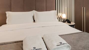 Una habitación de hotel moderna con una cama grande, dos almohadas, una mesita de noche con lámpara y dos toallas con el logotipo del hotel.