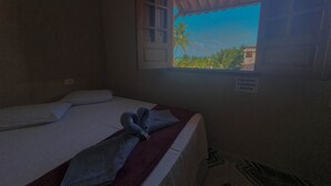 A single bed with a neatly folded towel and a window with a view of palm trees and a building. - Xaréu Flats Maragogi (Maragogi)