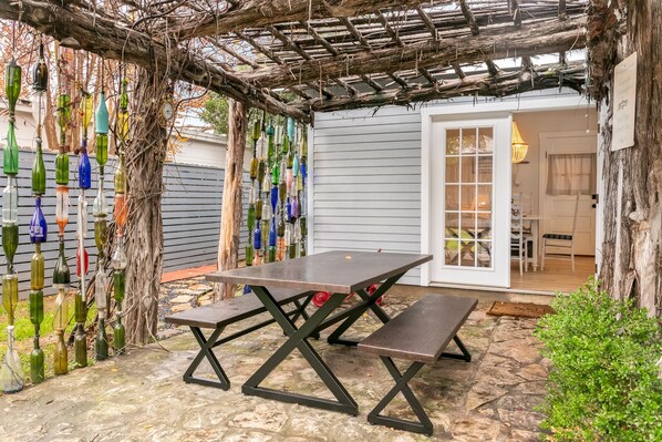 Un patio con una mesa y bancos, rodeado por una pérgola con botellas de vidrio colgantes.