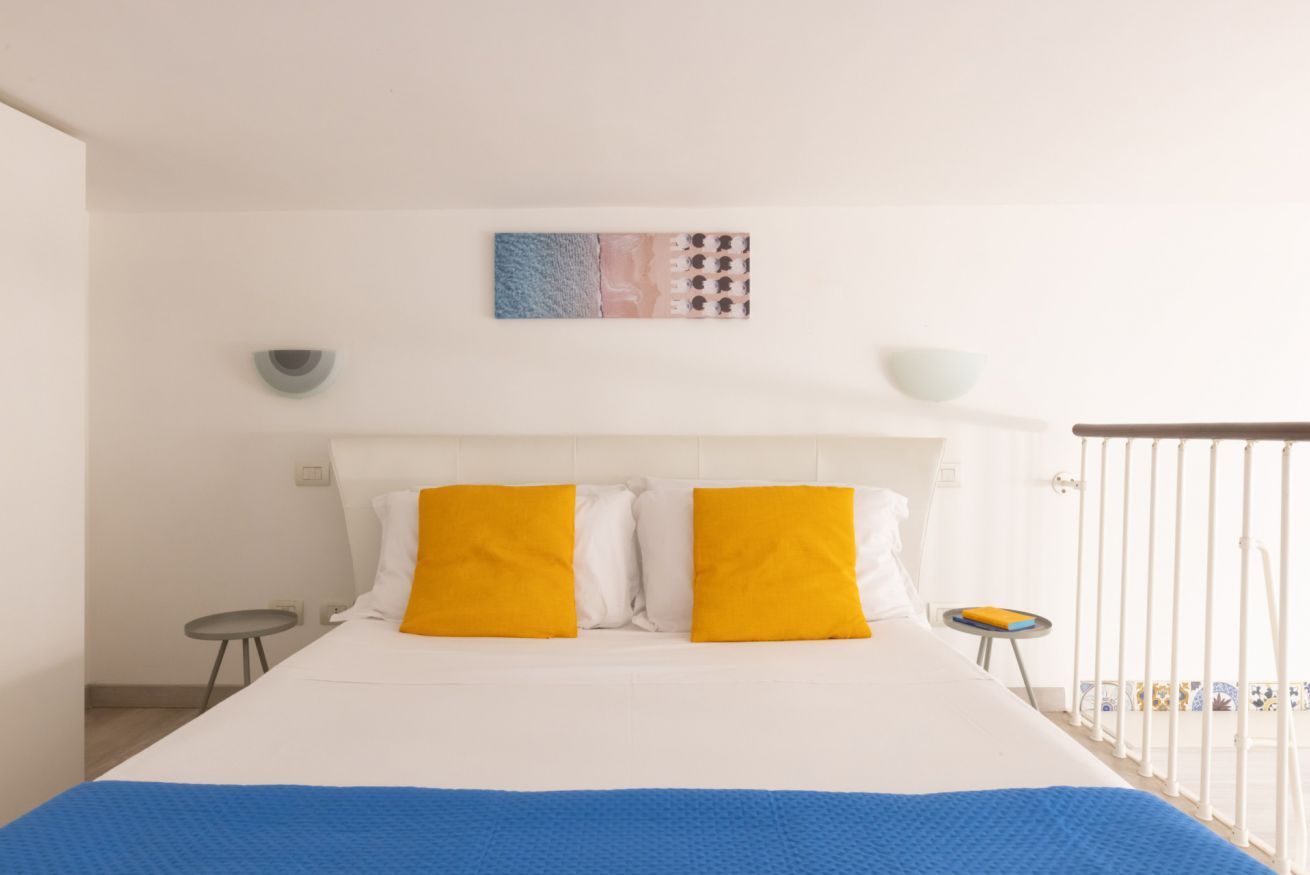 Un dormitorio con cama, dos almohadas amarillas, una colcha azul, una obra de arte en la pared, una mesita con un tazón y una barandilla blanca.