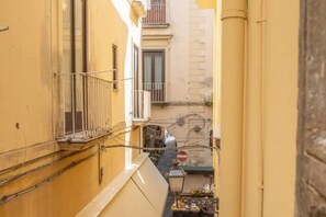 Une ruelle étroite bordée d’immeubles jaunes, avec un balcon bien visible et un petit étal de marché installé à l’extérieur.