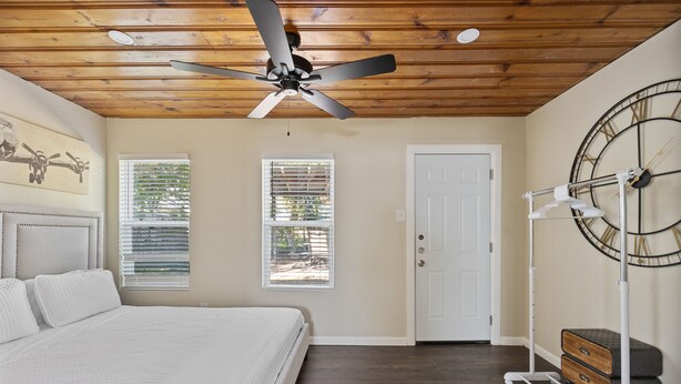 A bedroom with a bed, ceiling fan, two windows with blinds, a door, and a clock on the wall.