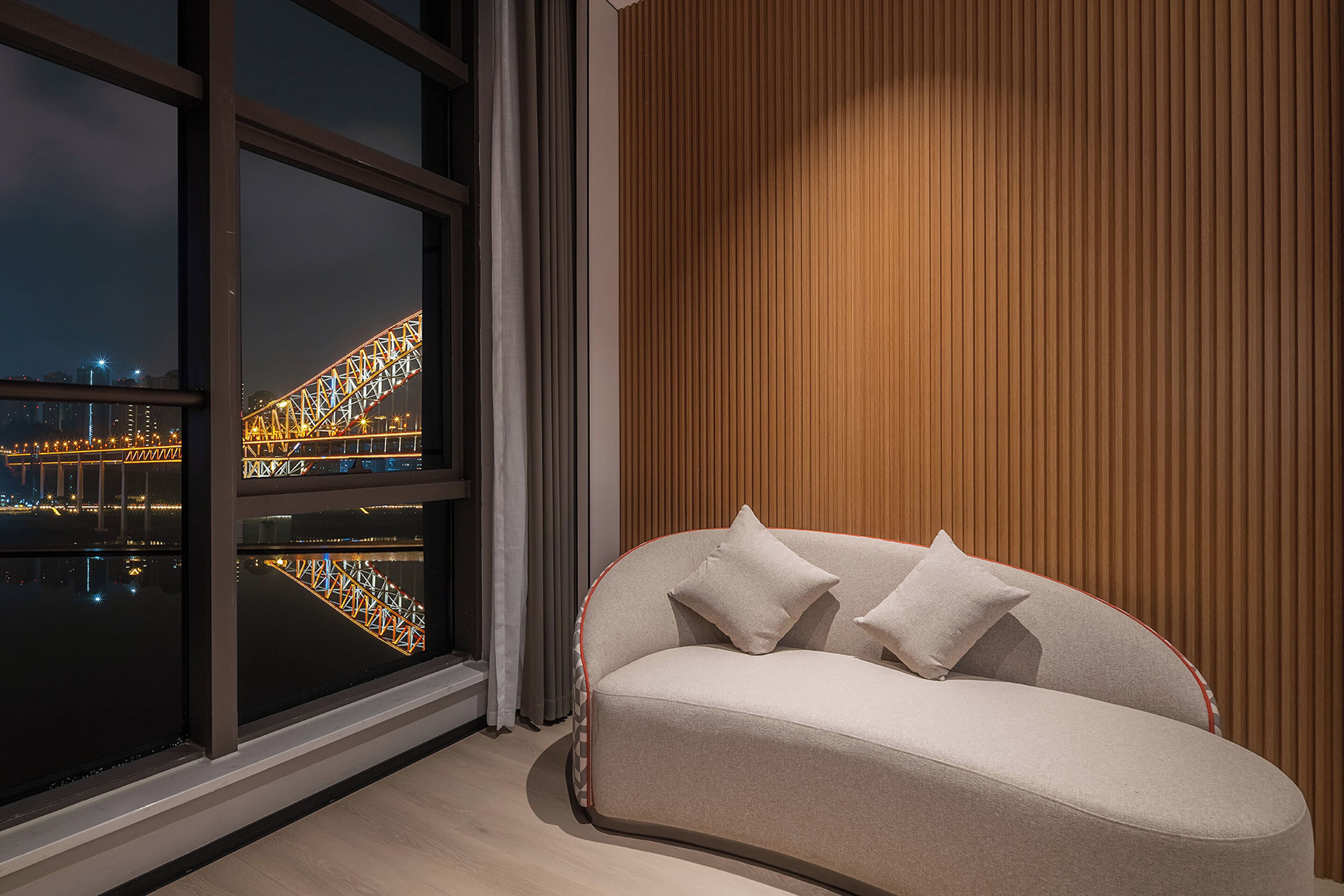 Ein modernes Wohnzimmer mit einer Couch, einem großen Fenster mit Blick auf eine beleuchtete Brücke und einer Holzwand.