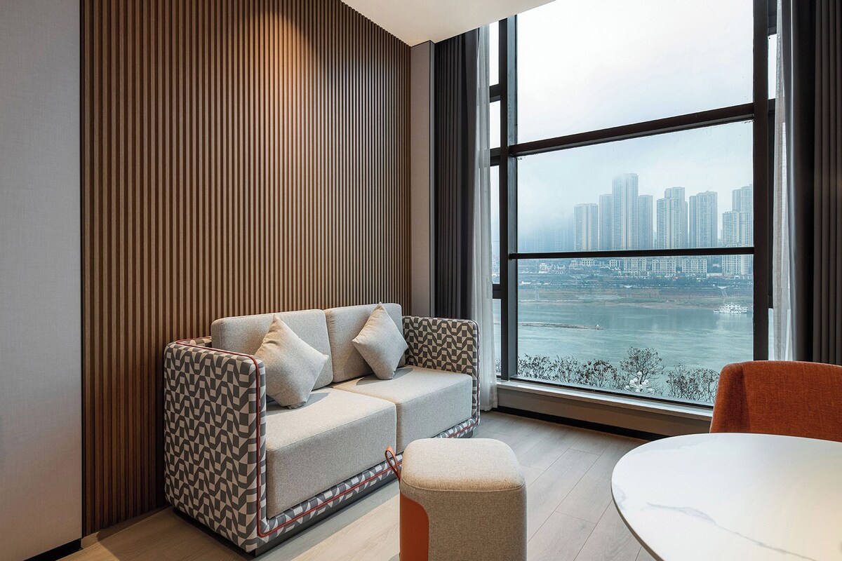A modern living room with a sofa, ottoman, and a table, featuring a large window with a city view.