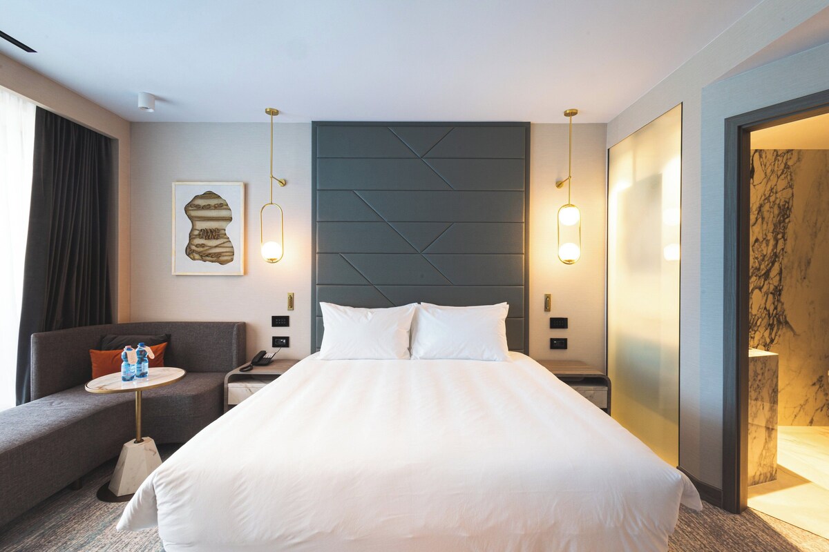 Ein Hotelzimmer mit einem großen Bett, einer Couch, einem kleinen Tisch mit Wasserflaschen und einem an der Wand befestigten Kunstwerk.