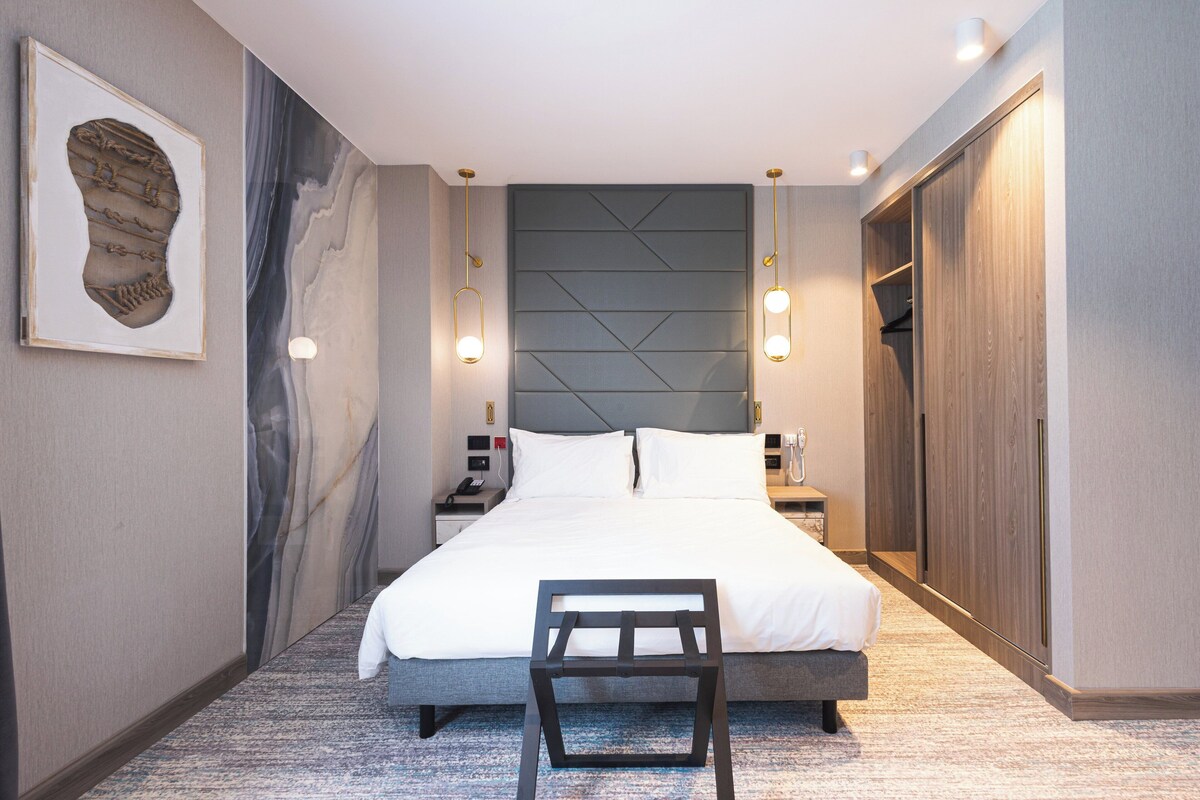 Ein modernes Hotelzimmer mit einem großen Bett, einem Nachttisch, einem Kleiderschrank und einem einzigartigen Kunstwerk an der Wand.