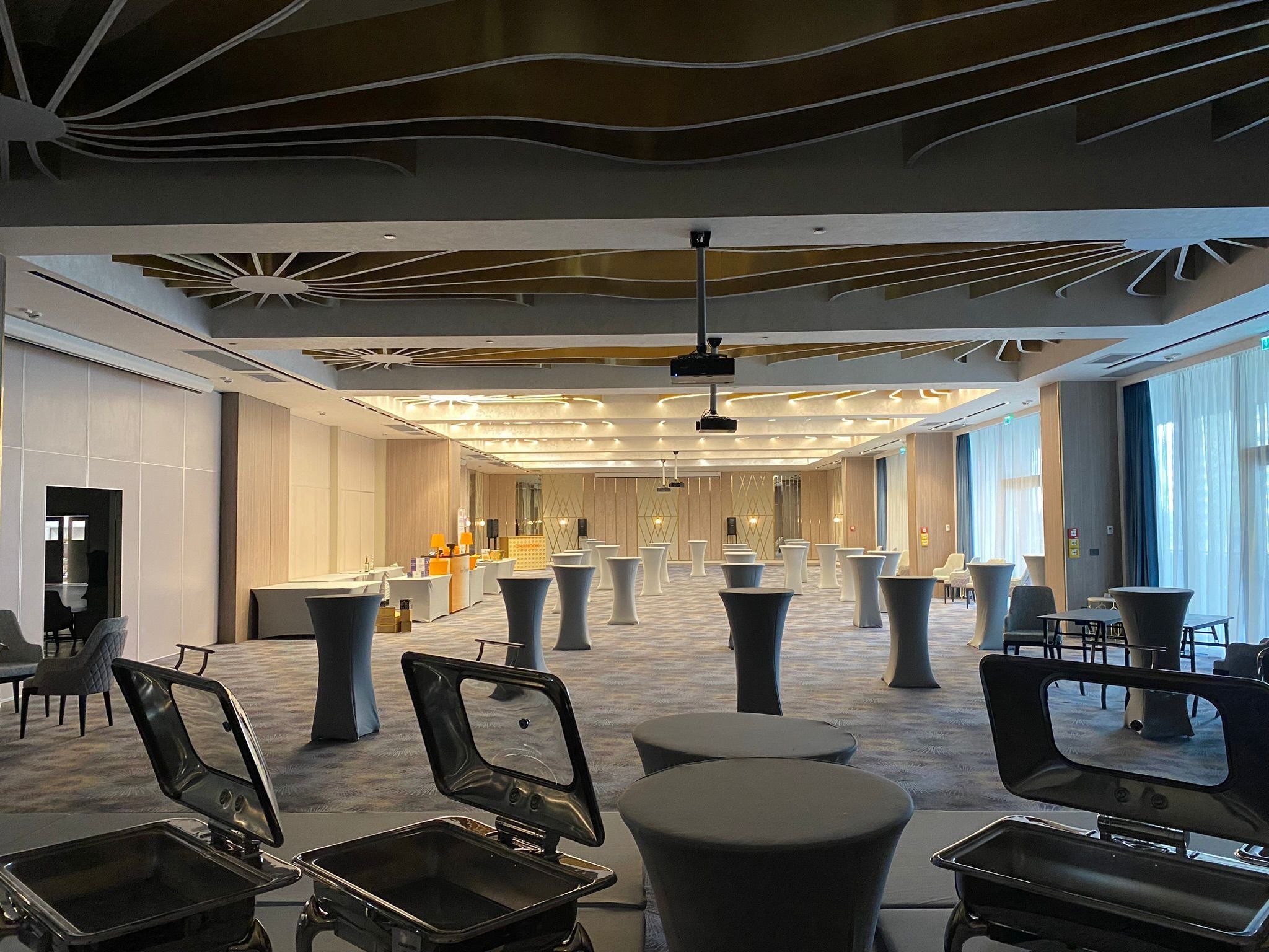 A spacious hotel lobby with a modern design, featuring a long reception desk, multiple seating areas, and a large window providing natural light.