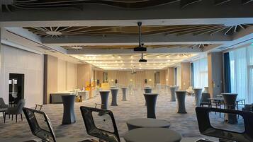 A spacious hotel lobby with a modern design, featuring a long reception desk, multiple seating areas, and a large window providing natural light.