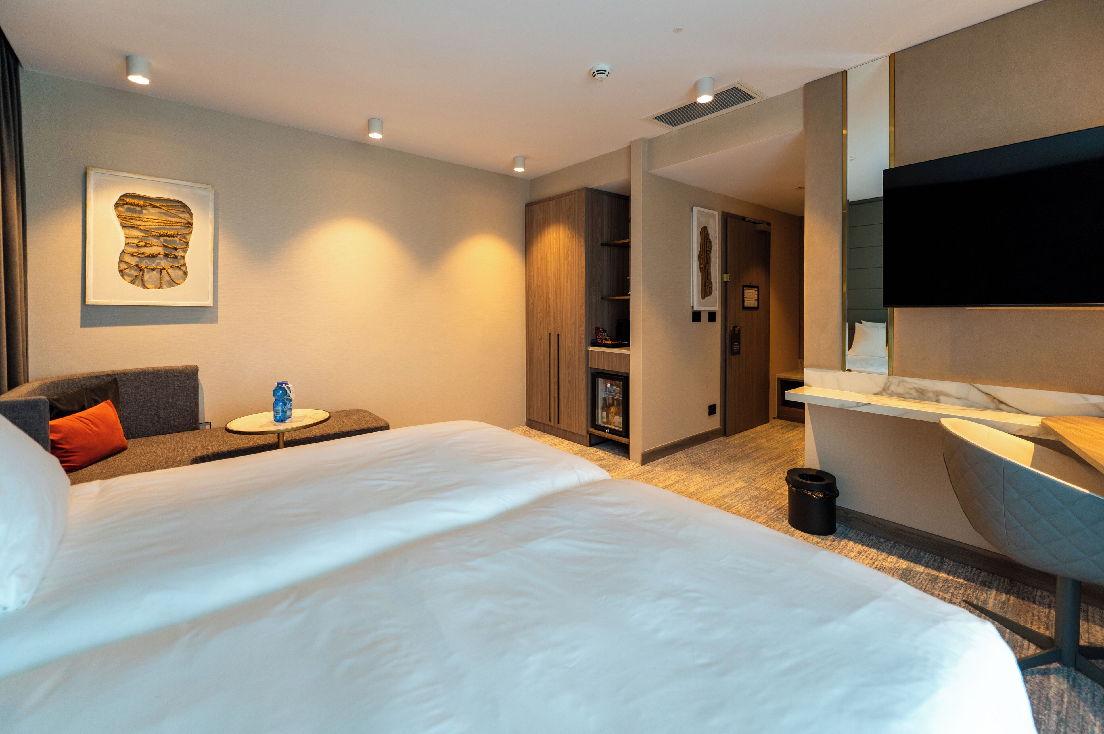 A modern hotel room with a large bed, a seating area with a small table, a minibar, a TV, and a bathroom with a mirror and sink.