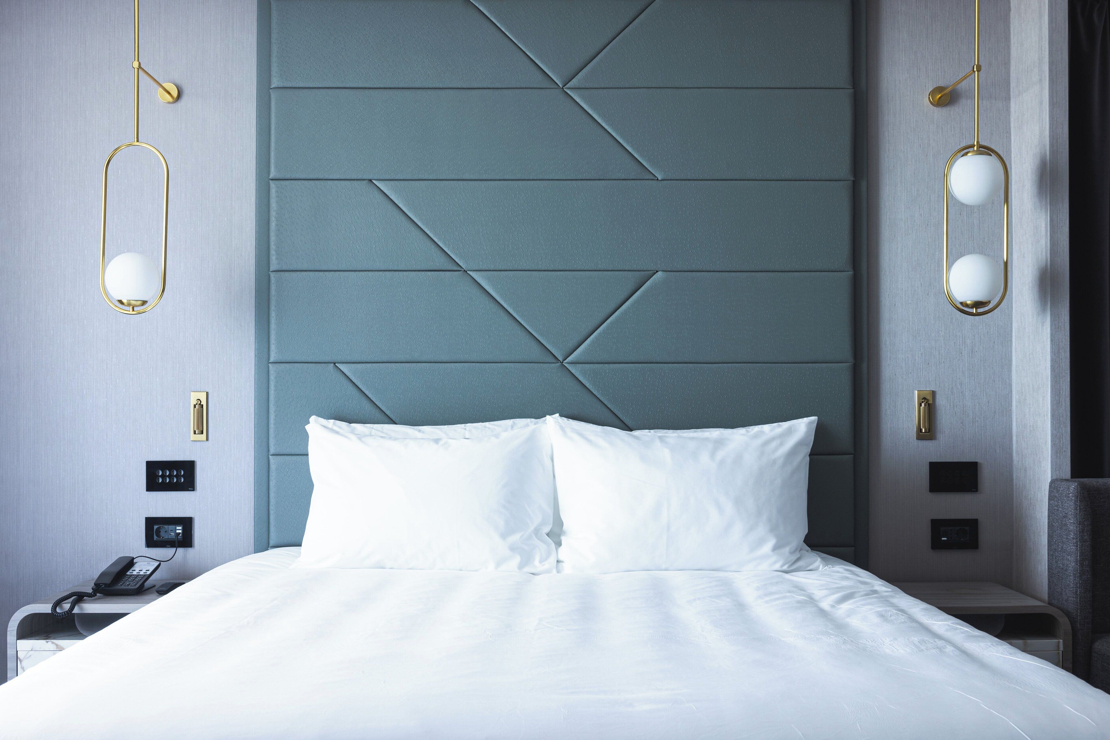 A neatly made bed with white linens, a modern headboard, bedside lamps, and a phone on the nightstand.