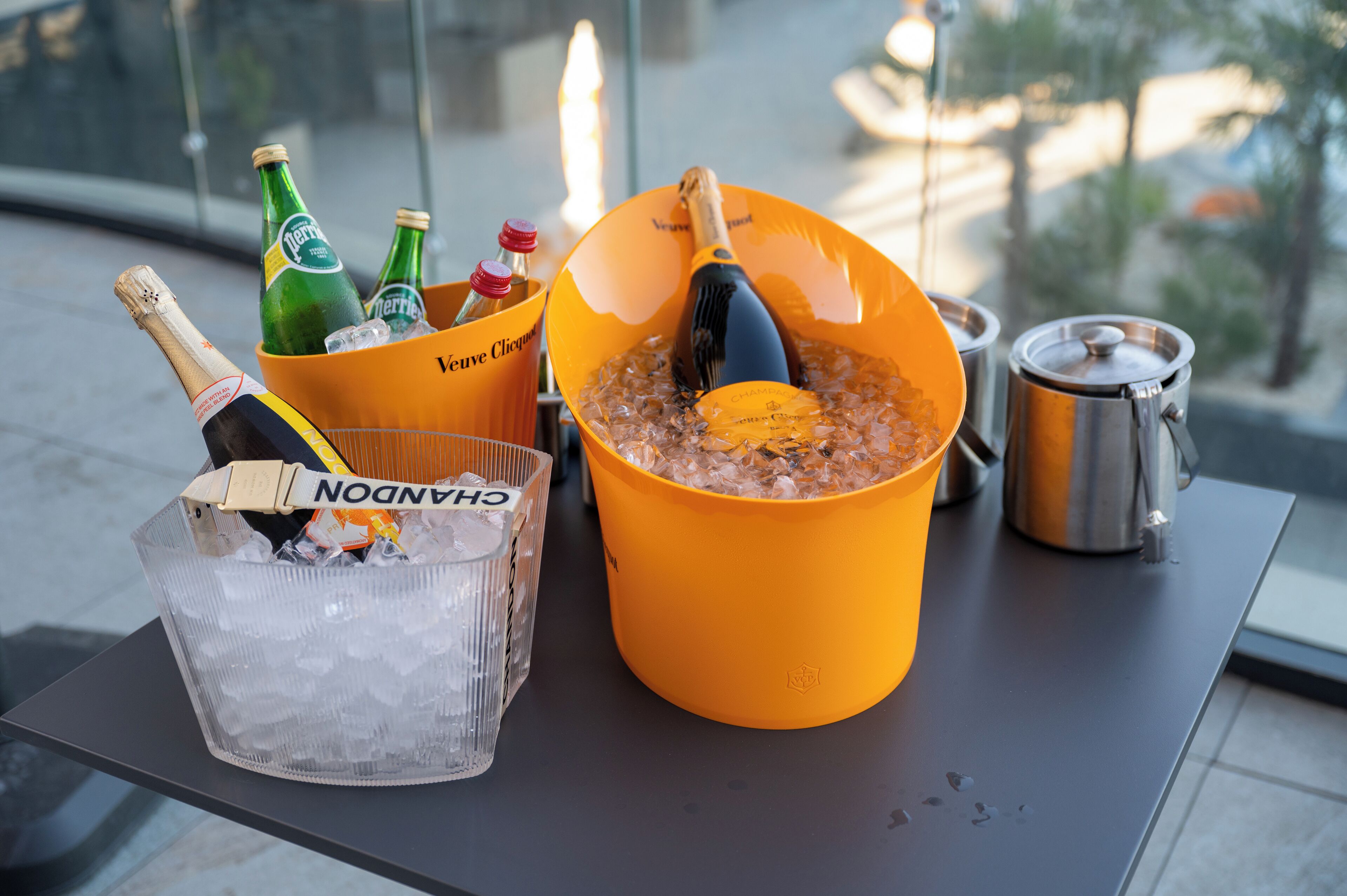 A table with a large yellow bucket containing a bottle of champagne, a bottle of Perrier, and a bottle of Cointreau. There is also a clear plastic bucket with ice and a bottle of Chandon champagne.
