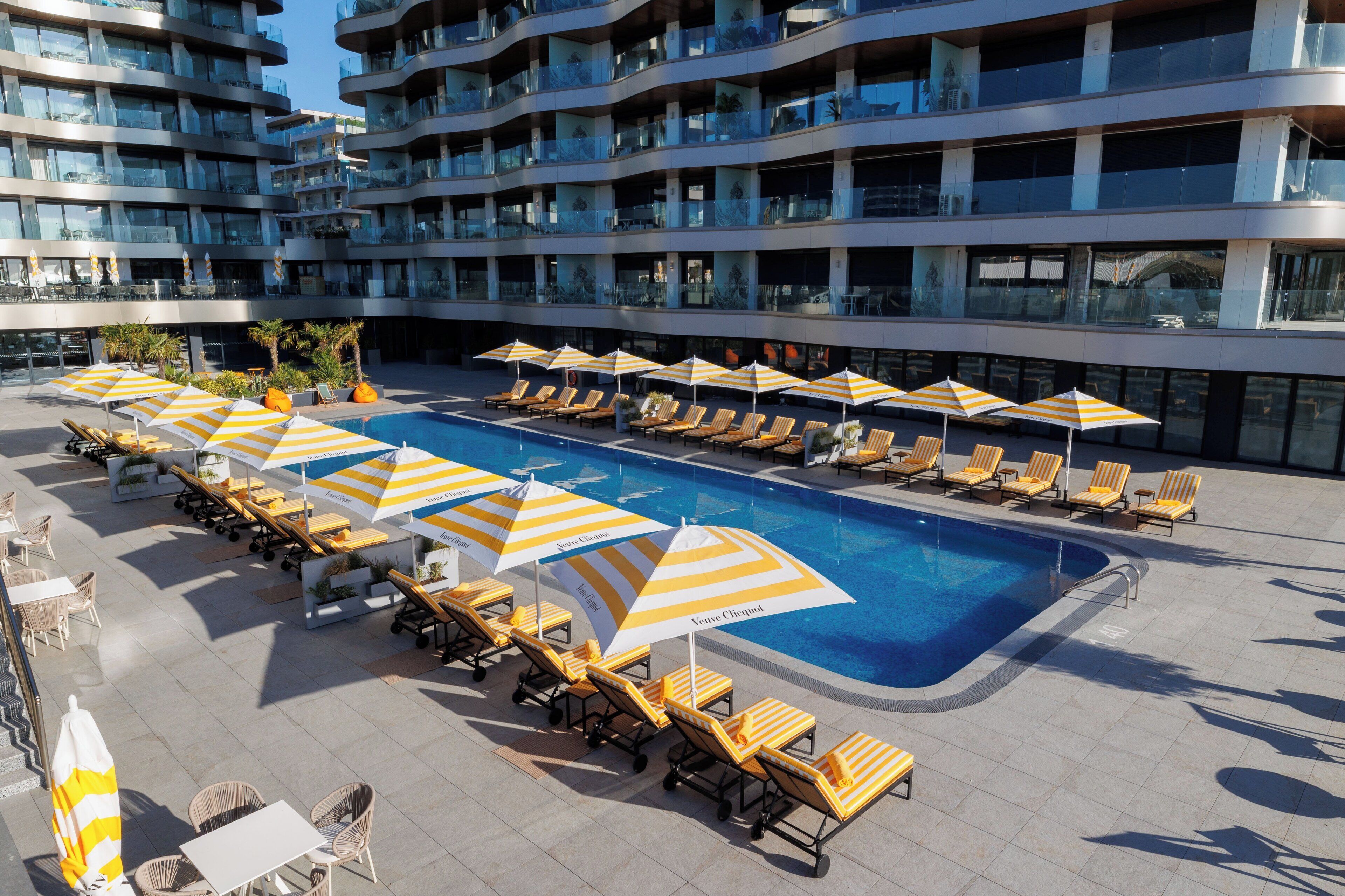 Un espace piscine aménagé sur le toit, avec des chaises longues et des parasols, entouré d’un immeuble moderne doté de balcons.