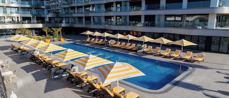 Un espace piscine aménagé sur le toit, avec des chaises longues et des parasols, entouré d’un immeuble moderne doté de balcons.