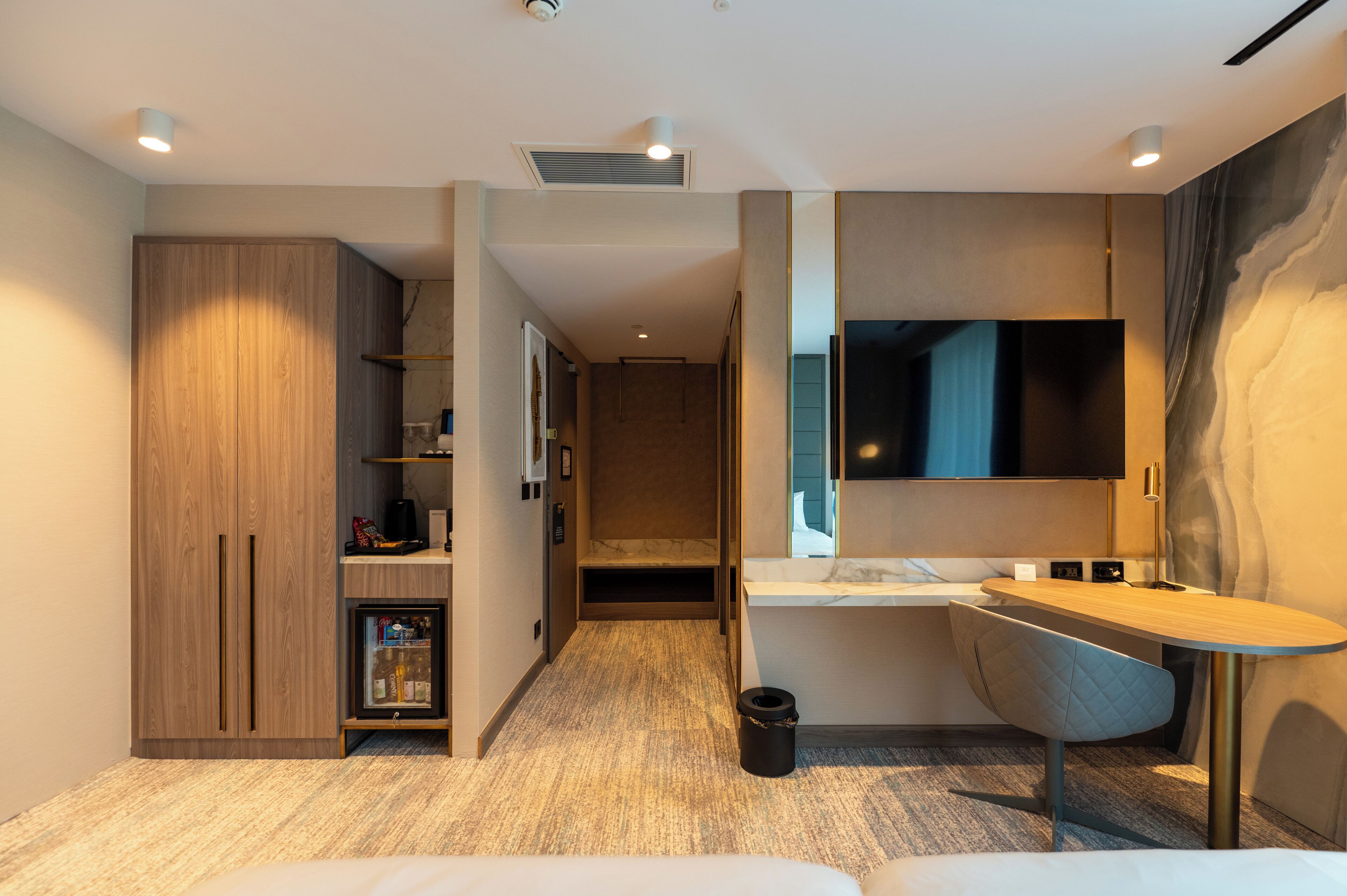 A modern hotel room with a wooden wardrobe, a built-in TV, a small desk, and a cozy seating area.