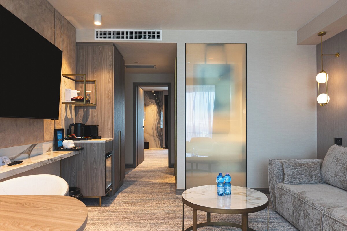 Ein modernes Hotelzimmer mit Küchenzeile, einer Sitzecke inklusive Sofa und rundem Tisch sowie einer großen Schiebeglasdoor zur Verbindung mit einem Flur.