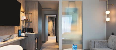 A modern hotel room with a kitchen area, a seating area with a sofa and a round table, and a large sliding glass door leading to a hallway.