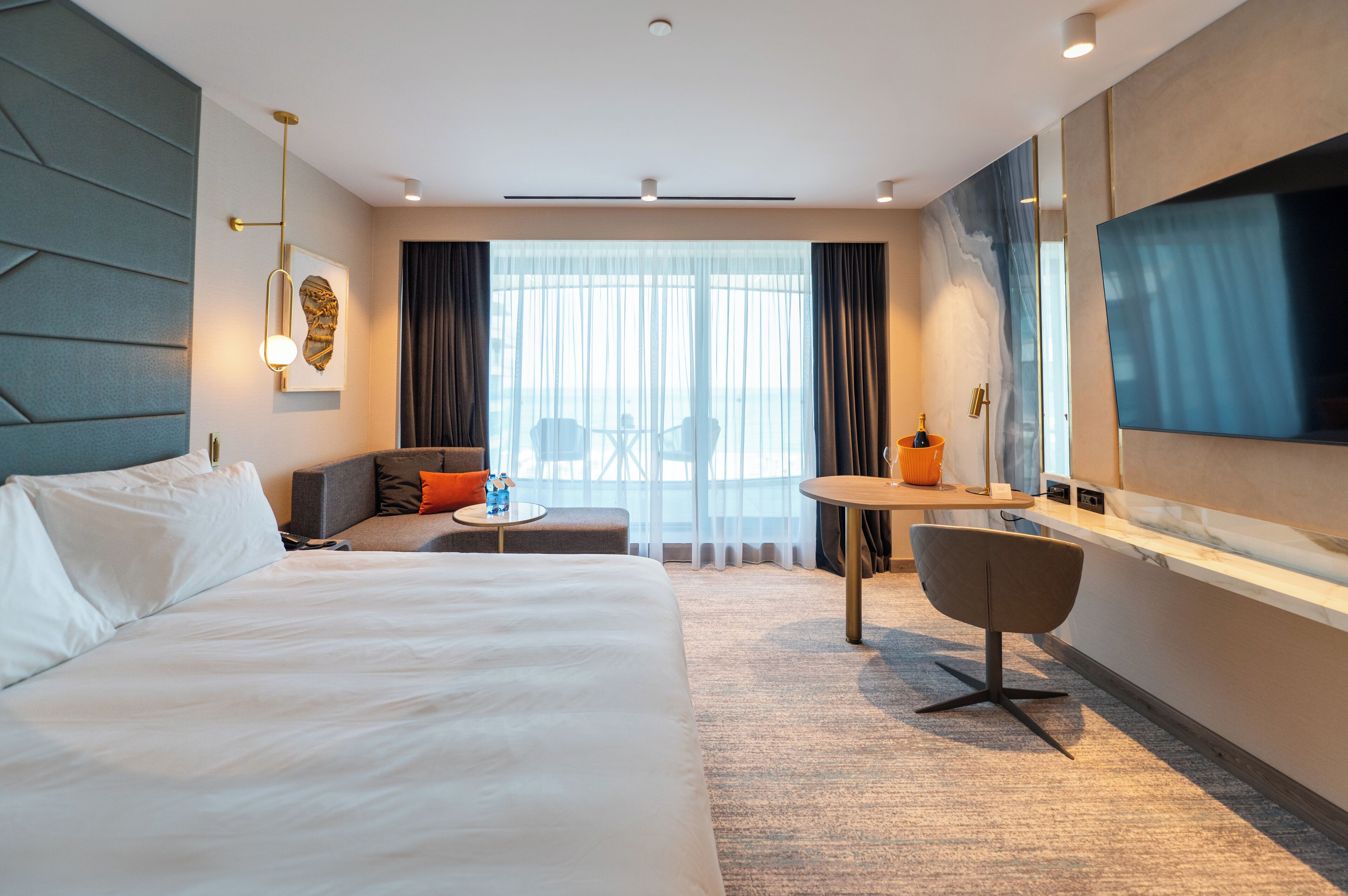 Una habitación de hotel moderna con una cama grande, un área de descanso con una mesita, un televisor y vistas a la ciudad a través de amplios ventanales.