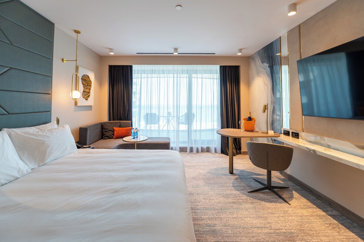 Ein modernes Hotelzimmer mit einem großen Bett, einer Sitzecke mit kleinem Tisch, einem Fernseher und Blick auf die Stadt durch große Fenster.