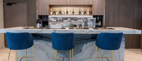 Un área de bar moderna con mostrador de mármol, tres taburetes azules y una estantería con botellas y copas.