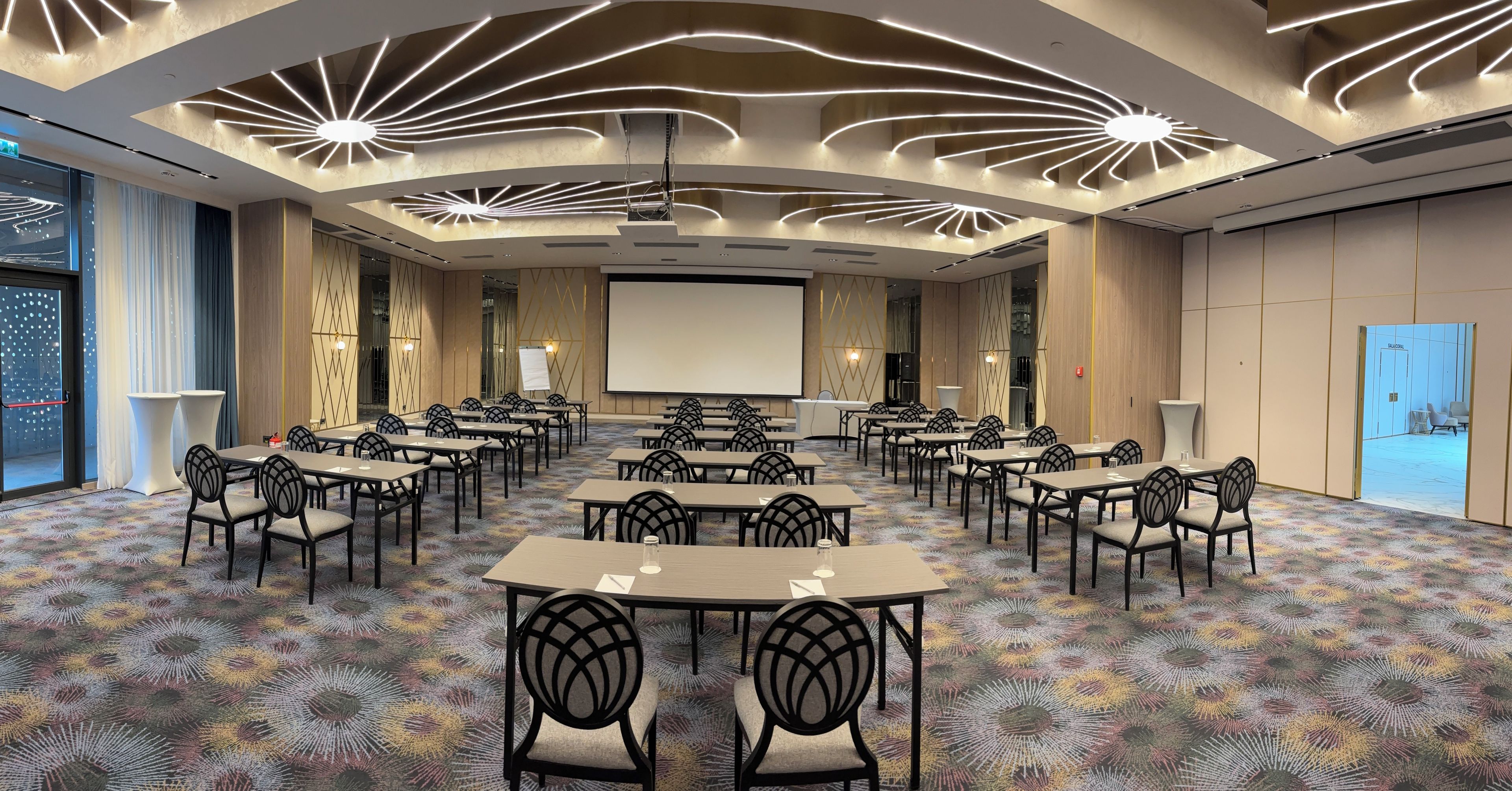 Un salón de conferencias con una pantalla grande, varias mesas y sillas dispuestas para reuniones.