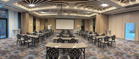Un salón de conferencias con una pantalla grande, varias mesas y sillas dispuestas para reuniones.