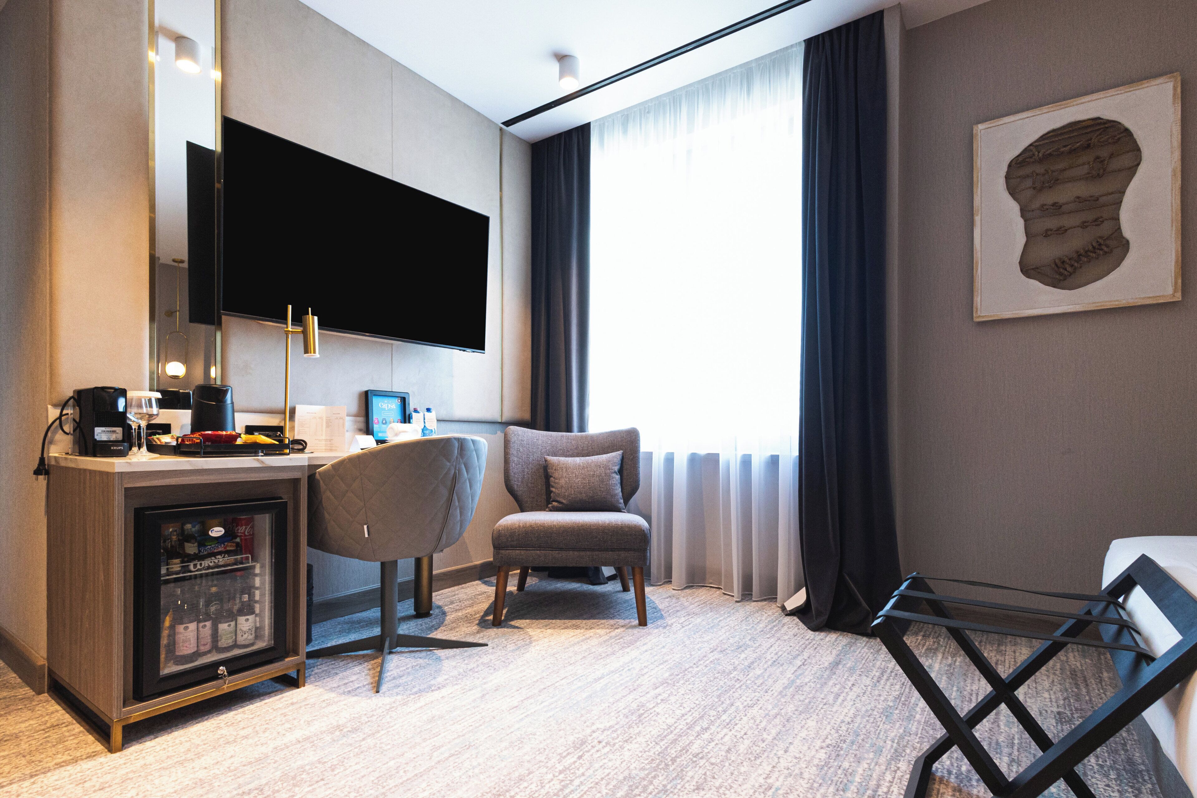 Habitación de hotel con televisión de pantalla plana montada en la pared, minibar, sillón cómodo, cama con estructura de metal negro y una ventana grande con cortinas.