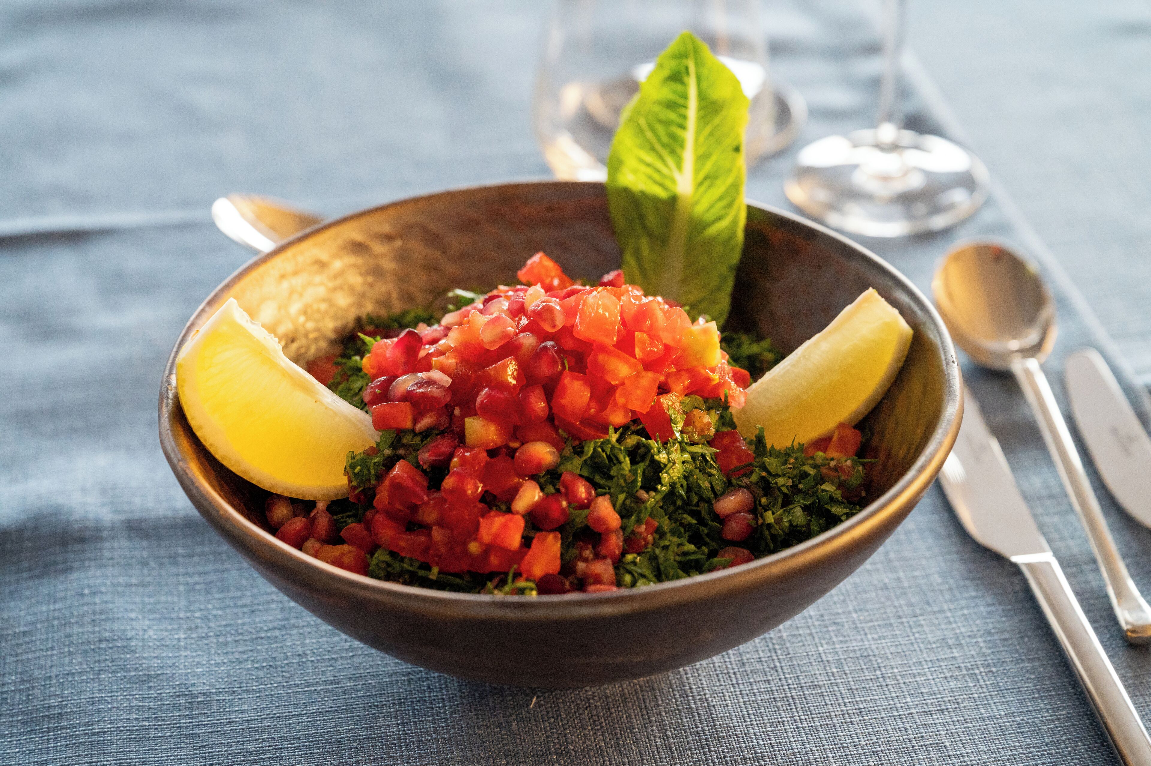 A bowl of salad with lemon wedges and a basil leaf on a blue tablecloth.