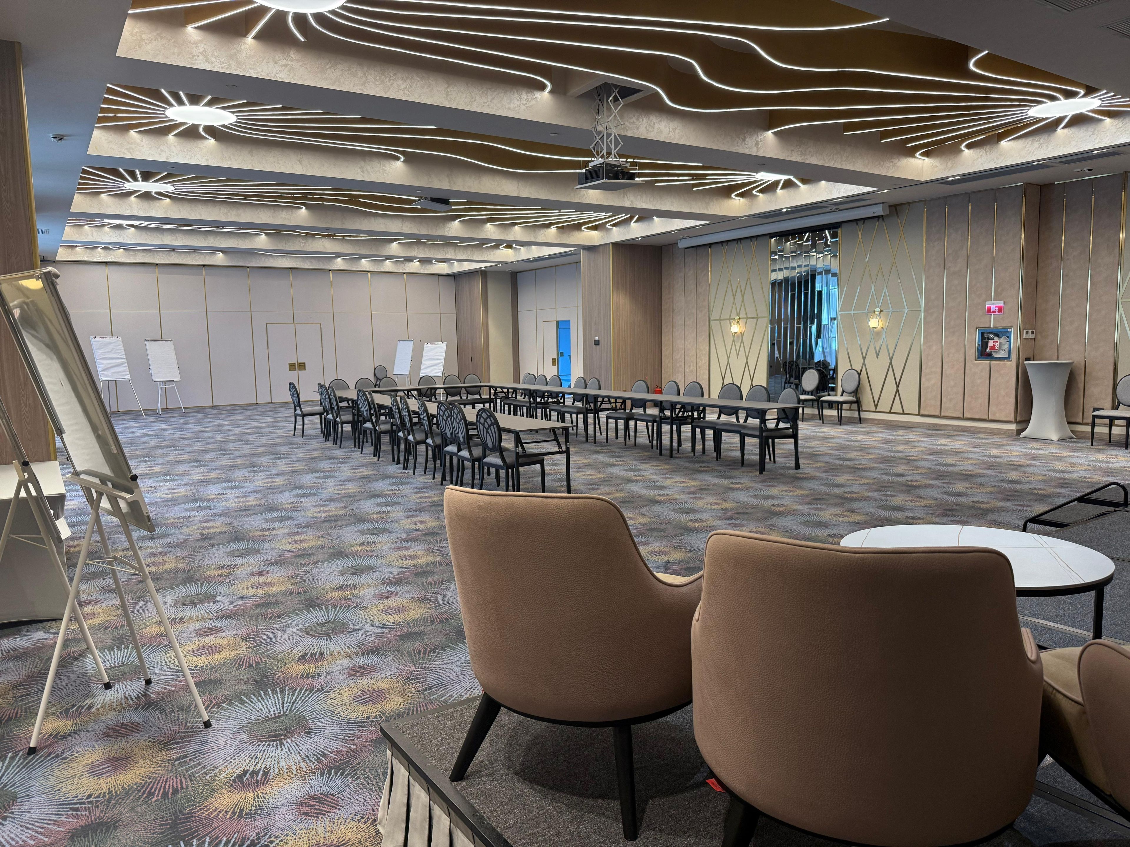 Egy konferenciaterem, melyben egy hosszú asztal, számos szék és szőnyeggel borított padló található.