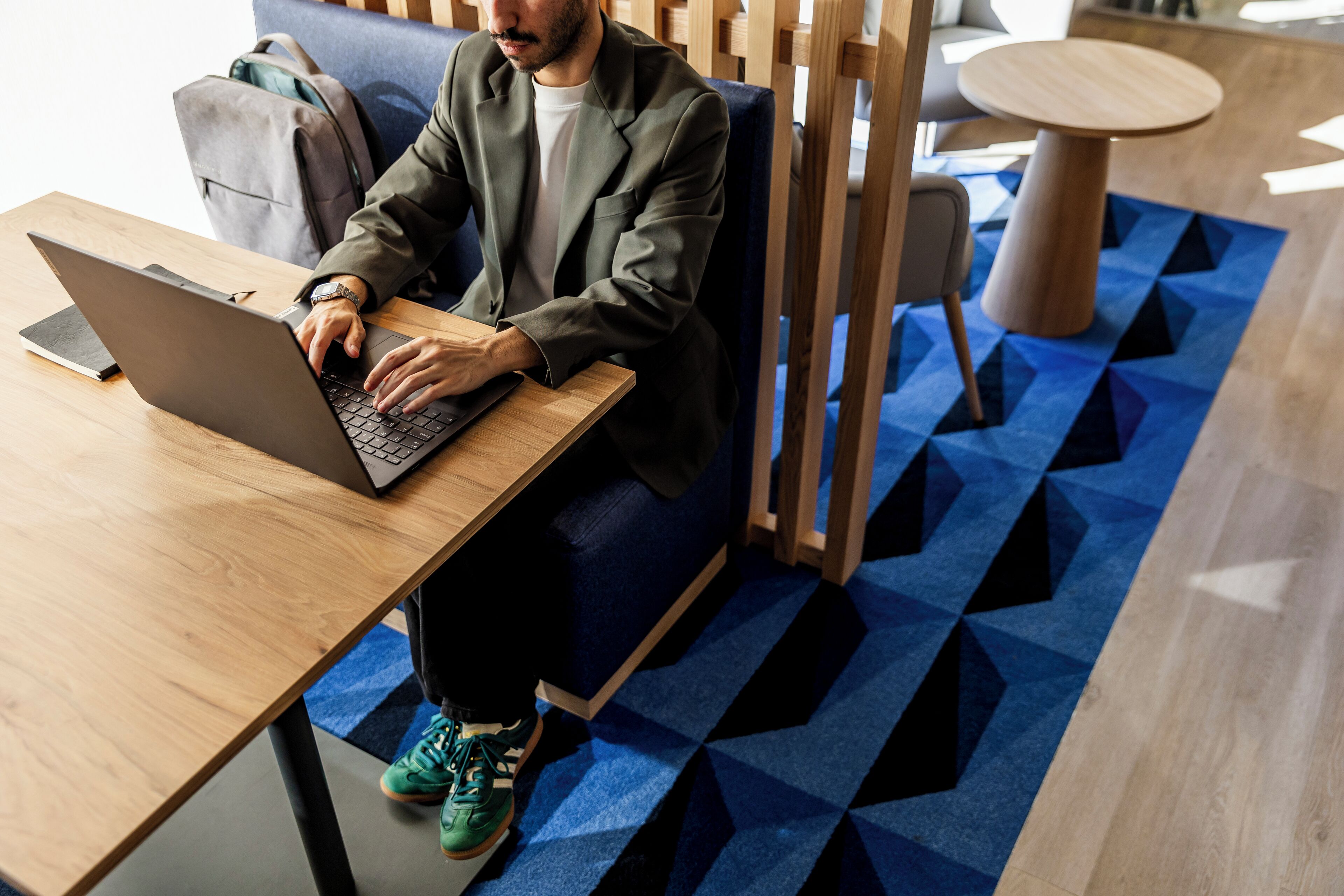 A person sitting at a wooden table with a laptop, in a room with a geometric patterned carpet and a round wooden table in the background.