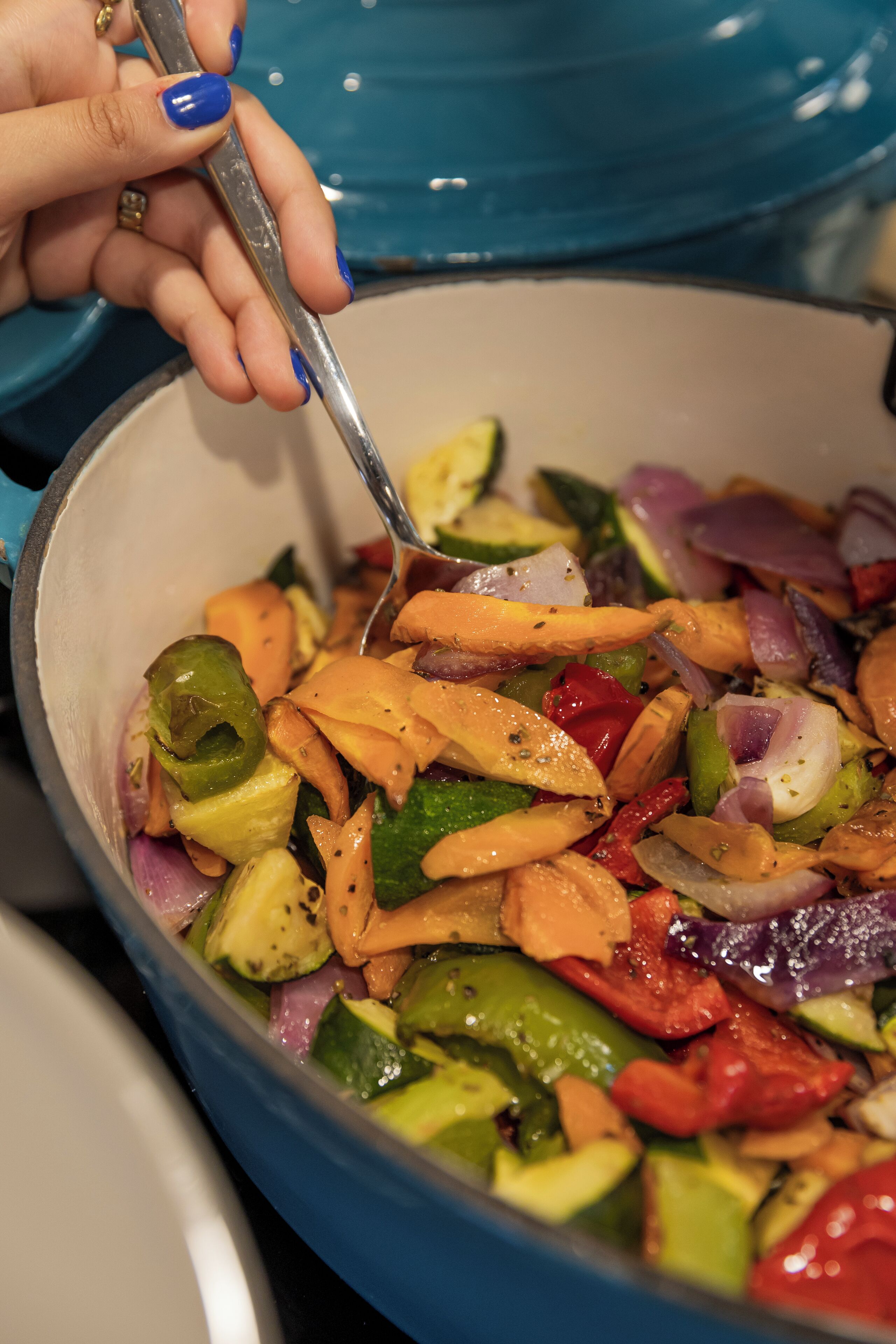 A close-up of a hand stirring a colorful vegetable dish in a blue pot.