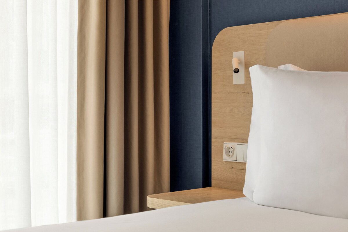 A hotel room with a wooden headboard, a white pillow, and a bedside table with a power outlet.