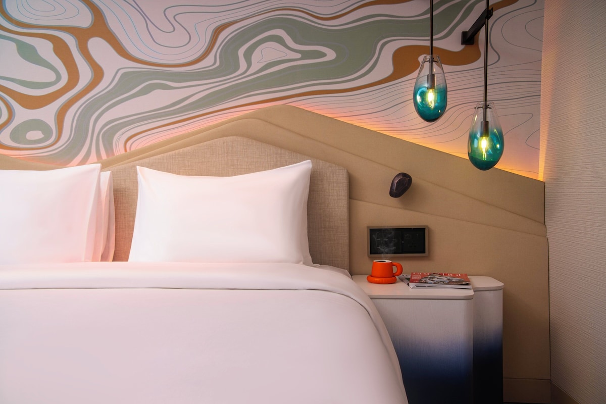 Ein modernes Hotelzimmer mit einem ordentlich bezogenen Bett, einem Nachttisch mit einer roten Tasse und einer Wand mit abstrakter Kunst.