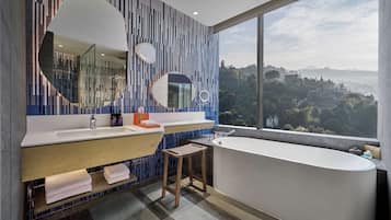 A modern bathroom with a large bathtub, two sinks, and a large mirror. There is a view of a mountainous landscape through a large window.