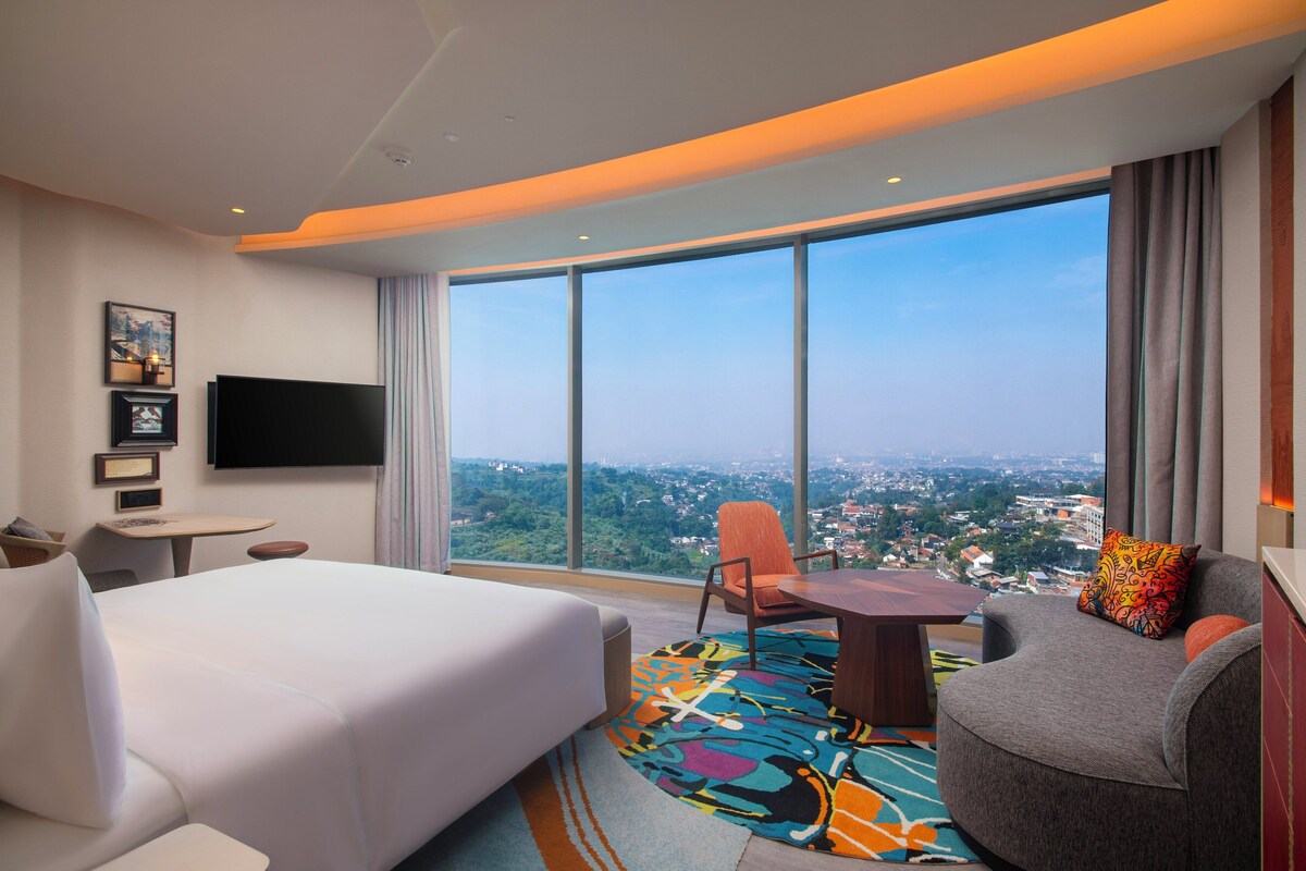Une chambre d’hôtel avec un grand lit, un bureau, une chaise et une télévision. Une grande fenêtre offre une vue sur la ville.
