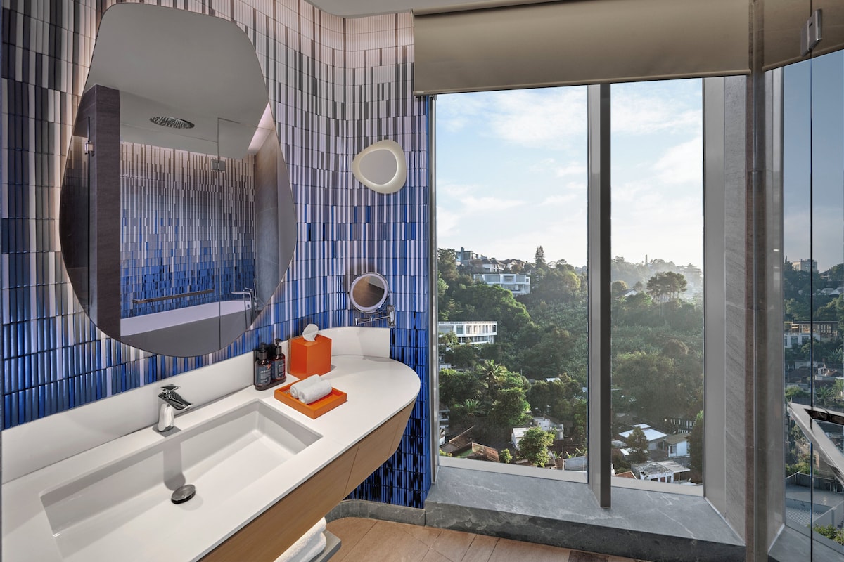 Ein modernes Badezimmer mit einem großen Spiegel, einem weißen Waschbecken und Blick auf eine Stadtlandschaft durch große Fenster.