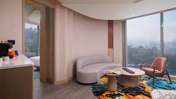 A modern hotel room with a curved wall, a large window offering a city view, a sofa, a chair, and a small table with a colorful rug.