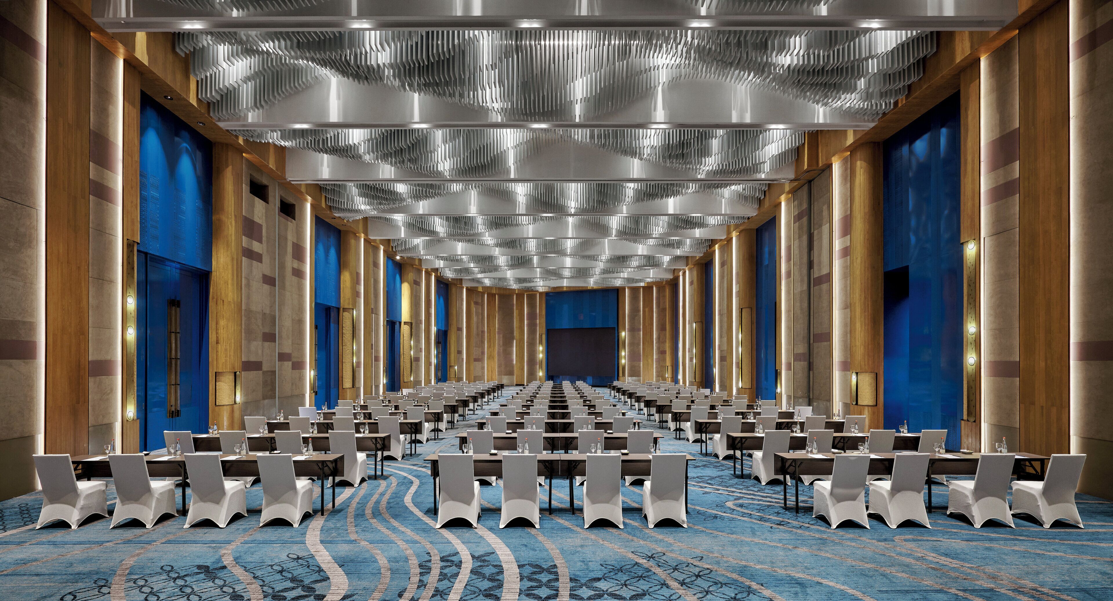 A large conference room with rows of tables and chairs, high ceilings with a unique light fixture, and a blue carpet with a pattern.