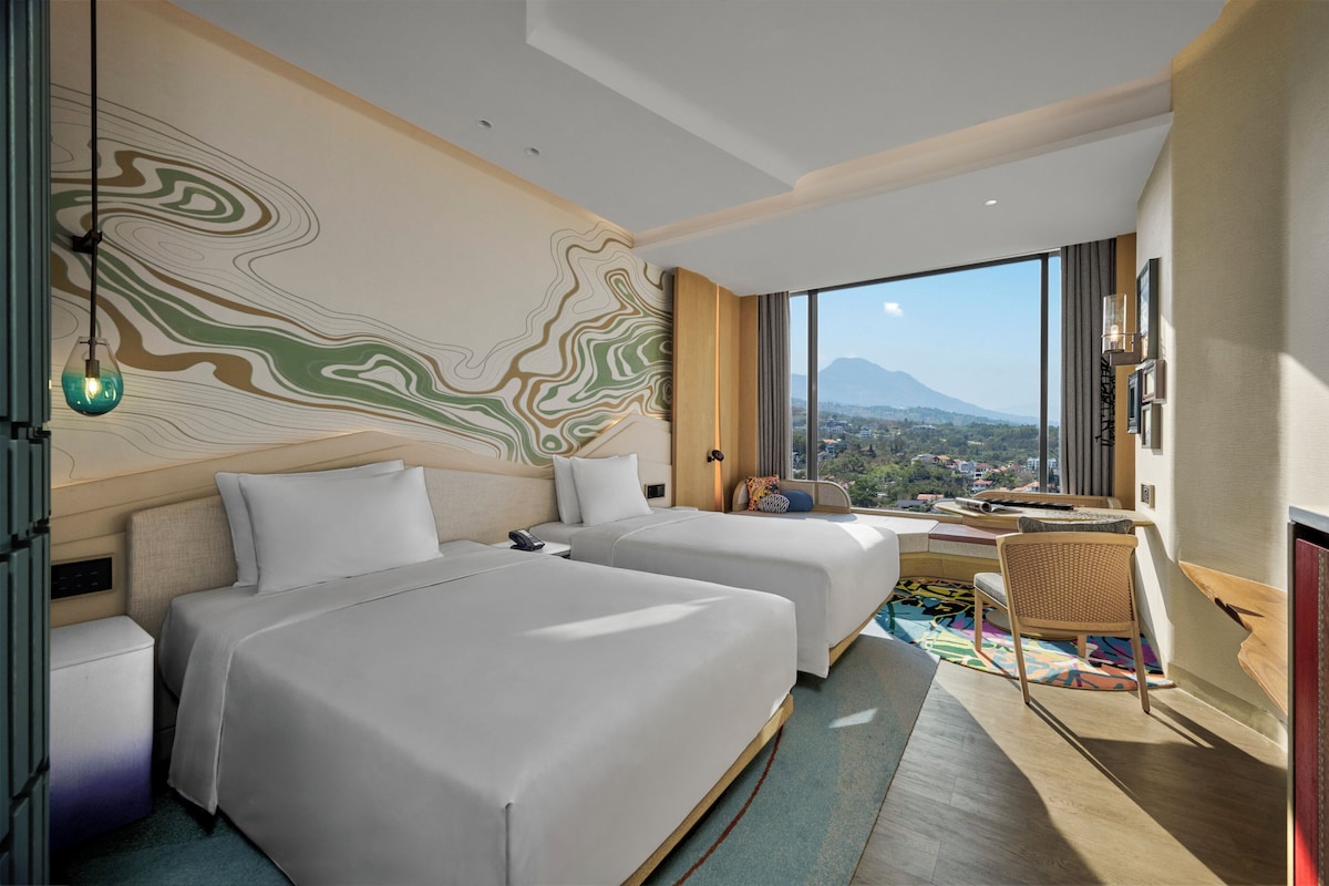 Ein Hotelzimmer mit zwei Betten, einem Schreibtisch, einem Sessel, einem Fernseher und einem großen Fenster mit Blick auf die Berge.