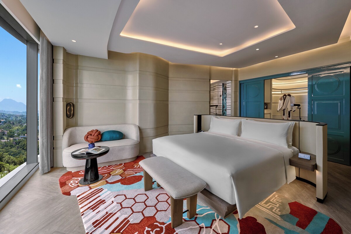 Une chambre d’hôtel moderne dotée d’un grand lit, d’un coin salon avec une petite table et offrant une vue sur le paysage urbain.