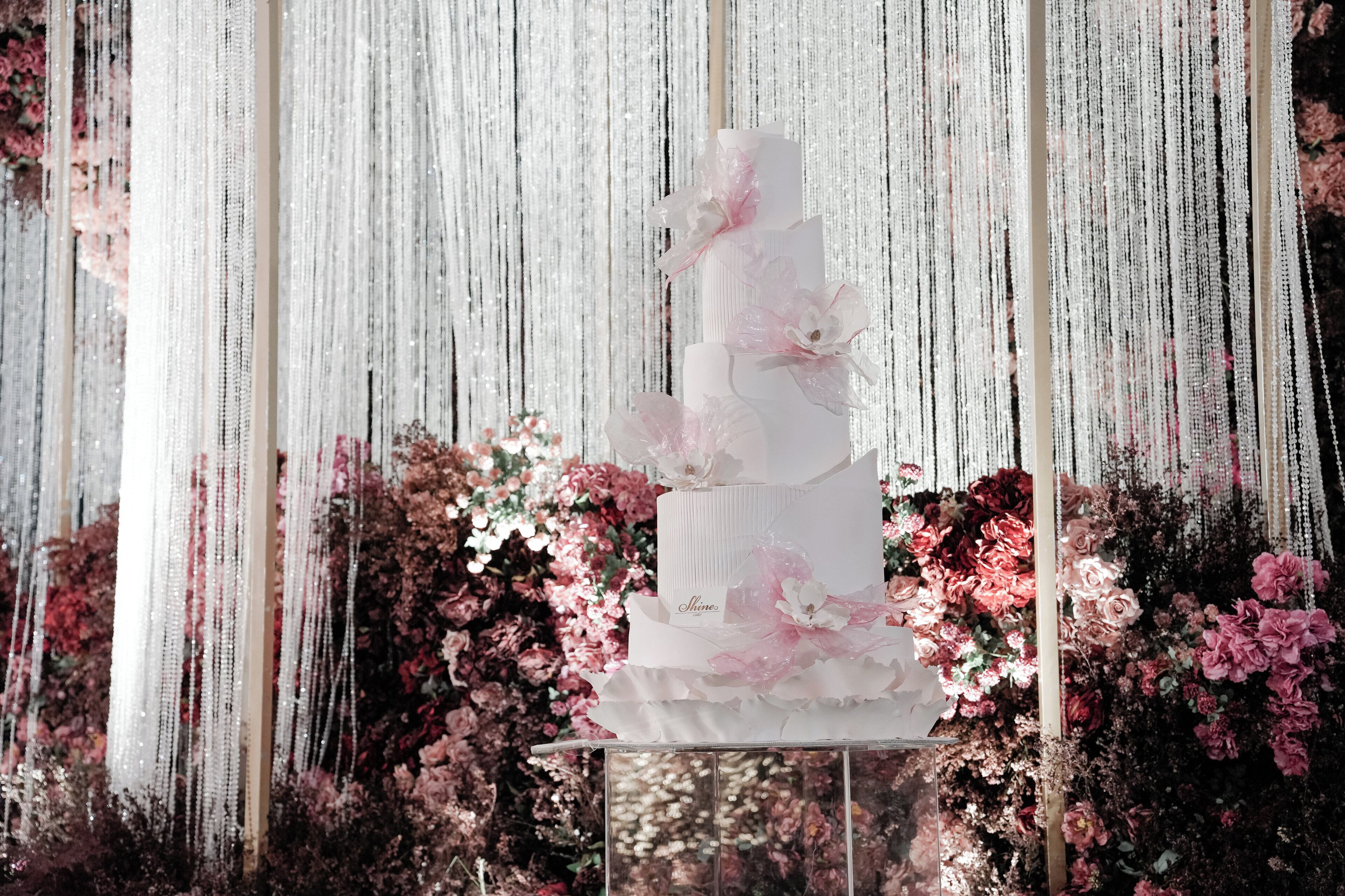 Un gâteau blanc à plusieurs étages, décoré de fleurs roses, est présenté sur un présentoir, avec en arrière-plan une guirlande de fleurs blanches et roses.